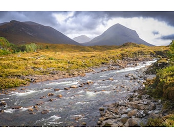 May include: A rocky river flows through a green valley with mountains in the background. The sky is cloudy and the water is flowing quickly.