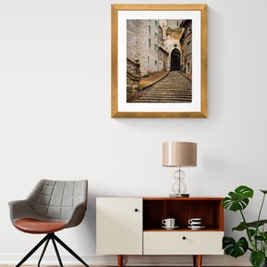 May include: A framed photograph of a narrow, cobblestone street in a European city. The street is lined with old buildings and leads up a set of stone steps to an archway.