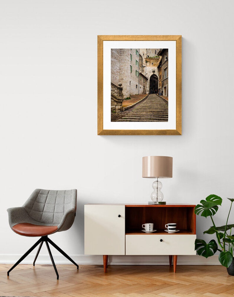 May include: A framed photograph of a narrow, cobblestone street in a European city. The street is lined with old buildings and leads up a set of stone steps to an archway.