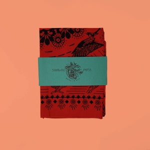 May include: A folded red bandana with black bird and floral designs. A teal paper band wraps around the bandana, with the text "SOURWOOD PRESS MADE IN NYC". The bandana is on a peach background.