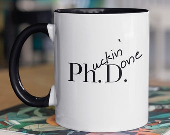 May include: White ceramic mug with a black rim and handle. The mug has black text that reads "Ph.D.  'uckin' Done". The mug is sitting on a stack of books with a floral pattern.