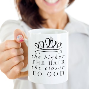 May include: White ceramic mug with a black crown design and the text "the higher the hair the closer to God". The mug is held up by a person. The mug is a standard size for coffee or tea.