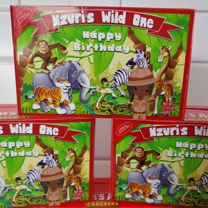 May include: A red box of animal crackers with a jungle theme. The box has a banner that says "Nzuri's Wild One Happy Birthday". The box features a cartoon image of a zebra, giraffe, elephant, tiger, and a girl dressed as an explorer. The box says "Net Wt 2.125 oz (60g)".