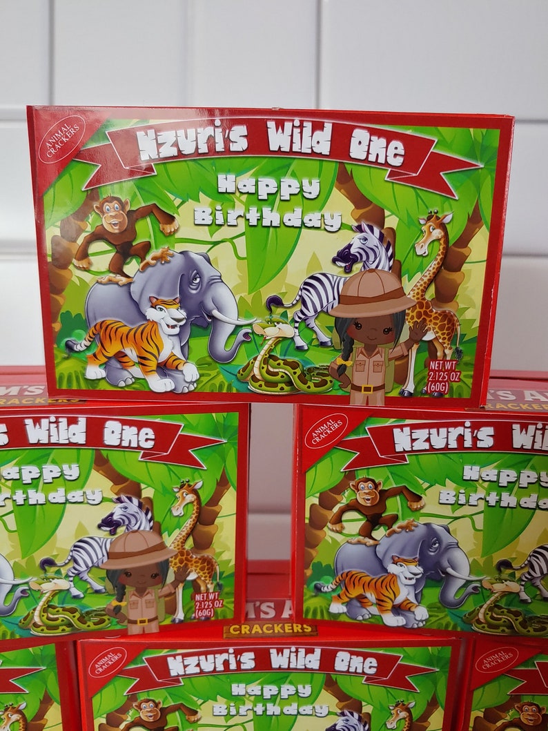 May include: A red box of animal crackers with a jungle theme. The box has a banner that says "Nzuri's Wild One Happy Birthday". The box features a cartoon image of a zebra, giraffe, elephant, tiger, and a girl dressed as an explorer. The box says "Net Wt 2.125 oz (60g)".
