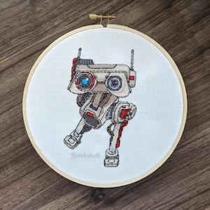 May include: Cross-stitch embroidery of a robot character in a wooden hoop. The robot is primarily beige, with red and black accents. It has two large eye-like features, one with a blue center. The word "Bombshell" is stitched at the bottom.