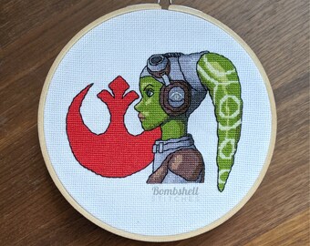 May include: Cross-stitch embroidery of a green-skinned character with a red Rebel Alliance symbol in the background. The character has a grey helmet and green hair. The text "Bombshell Stitches" is at the bottom of the embroidery.