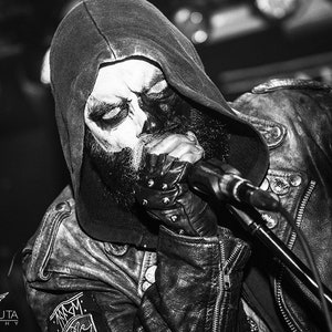 May include: A black and white photo of a person wearing a black hooded jacket with a skull face paint and a microphone in front of their mouth.
