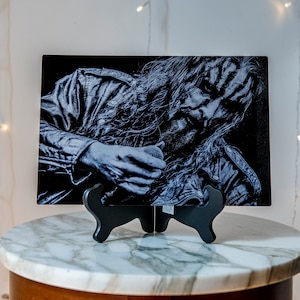 May include: Black and white metal print of a person with long hair and a leather jacket. The print is on a black stand and is displayed on a marble surface.