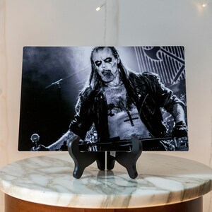 May include: Black and white photo of a musician with long hair and a beard, wearing a leather jacket and a t-shirt with a cross on it. The photo is printed on a metal sheet and displayed on a black stand.