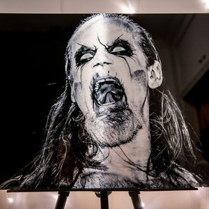 May include: Black and white portrait of a man with long hair and a beard, screaming with his mouth open and tongue out. He is wearing black eye makeup and has a dark background.