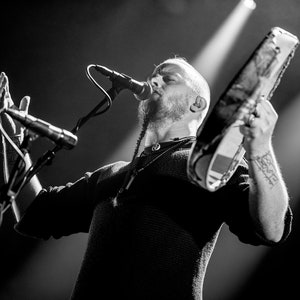 May include: A man with a beard and a long braid sings into a microphone while holding a tambourine. He is wearing a black sweater and has a tattoo on his arm.