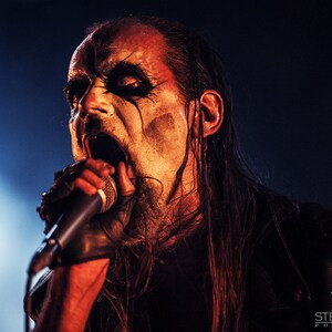 May include: A man with long black hair and a black and white face paint sings into a microphone. He is wearing a black shirt and has a microphone in his hand.