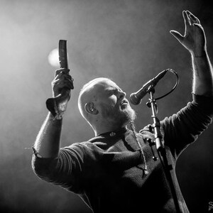 May include: A man with a shaved head sings into a microphone while holding a wooden instrument. He is wearing a dark sweater and has a beard.