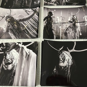 May include: Black and white photograph of a person wearing a headdress with antlers and a long, flowing garment. The person is singing into a microphone. The background is blurred and out of focus.