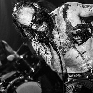 May include: A black and white photo of a man with long black hair and a beard, wearing a black leather glove and a necklace with a pendant. He has several tattoos on his chest and arms, including a cross and a map. He is singing into a microphone.