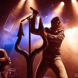 May include: A person with long hair sings into a microphone while holding a large black trident. The person is wearing a black leather jacket and blue jeans. The stage is lit with bright lights.