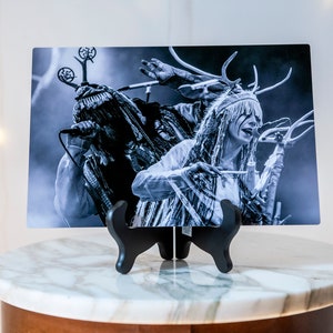 May include: Black and white photo of two people wearing elaborate costumes with antlers and feathers, performing on stage. The photo is printed on a metal sheet and displayed on a black stand.