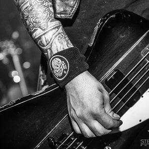 May include: A black and white photo of a person playing a black electric bass guitar. The guitar has a white pickguard with the word "HellYeah" written on it. The person is wearing a black wristband with a skull and crossbones design.