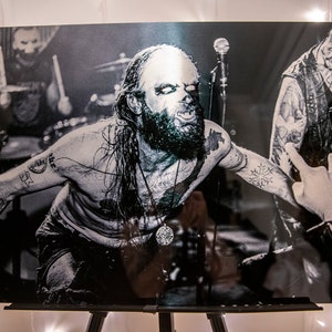 May include: Black and white photograph of a man with long hair and a beard performing on stage. He is wearing a necklace with a large pendant and has tattoos on his arms. The man is looking intensely at the camera.