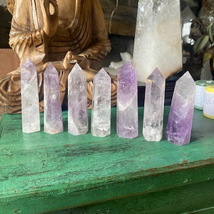 May include: Several amethyst crystal points, varying in colour from clear to deep purple, are displayed on a distressed green wooden surface. A large clear quartz crystal and a wooden Buddha statue are in the background.