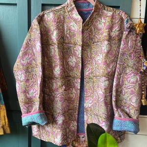 May include: A long-sleeved quilted jacket featuring a tan base with a repeating pink floral pattern. The garment has a mandarin collar, a straight front opening, and cuffs that are folded back to reveal a contrasting blue and pink interior lining.