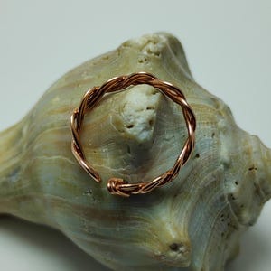 May include: A copper-colored, adjustable ring with a twisted rope design. The ring is open at one end and sits on a light green and tan seashell. The ring's design features a series of intertwined wires, creating a textured appearance.
