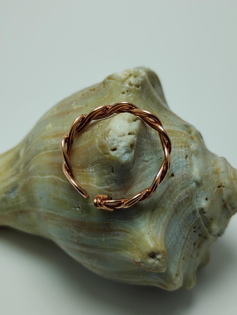 May include: A copper-colored, adjustable ring with a twisted rope design. The ring is open at one end and sits on a light green and tan seashell. The ring's design features a series of intertwined wires, creating a textured appearance.
