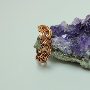 May include: A copper-colored ring with an intricate woven design. The ring is displayed on a rough, gray rock with a cluster of purple amethyst crystals. The background is a soft, off-white color.
