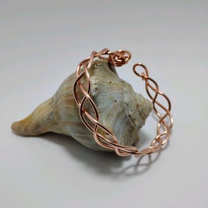 May include: A copper-colored bracelet with an open design, resting on a seashell. The bracelet features an intricate, interwoven pattern. The seashell is tan and has a smooth texture. The background is a soft, neutral color.