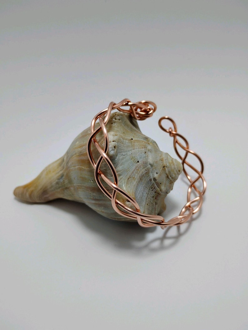 May include: A copper-colored bracelet with an open design, resting on a seashell. The bracelet features an intricate, interwoven pattern. The seashell is tan and has a smooth texture. The background is a soft, neutral color.
