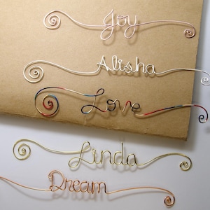 May include: Five wire bookmarks with different names written on them. The names are Joy, Alisha, Love, Linda, and Dream. The bookmarks are made of different colored wires, including gold, silver, and red.