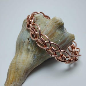 May include: A copper-colored wire bracelet with a woven design rests on a light brown seashell. The bracelet's links are formed by wrapping the wire, creating a textured, interconnected pattern. The seashell provides a natural backdrop.