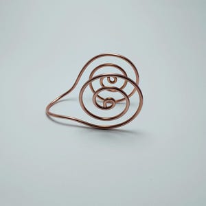 K&ouml;nnte beinhalten: Ein kupferfarbenes Drahtarmband mit einem Spiraldesign. Das Armband hat konzentrische Kreise und ein gebogenes Band. Das Metall hat eine polierte Oberfl&auml;che, die das Licht reflektiert. Das Design ist minimalistisch und modern.