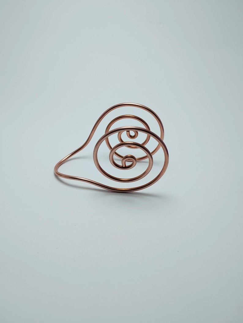K&ouml;nnte beinhalten: Ein kupferfarbenes Drahtarmband mit einem Spiraldesign. Das Armband hat konzentrische Kreise und ein gebogenes Band. Das Metall hat eine polierte Oberfl&auml;che, die das Licht reflektiert. Das Design ist minimalistisch und modern.