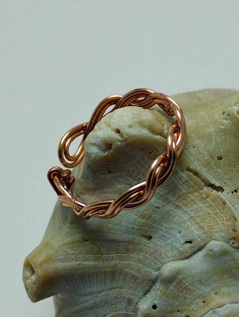 May include: A copper-colored ring with a braided design. The ring is made of twisted wire, forming a continuous loop. The ring is resting on a light-colored, textured surface, possibly a seashell. The ring's design is simple yet elegant.