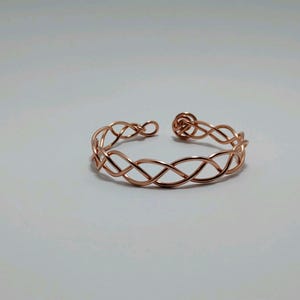 May include: A copper-colored open cuff bracelet with an intricate woven design. The bracelet features a series of intertwined loops, creating a delicate and stylish accessory. The open design allows for easy wear and adjustment. Jewelry.