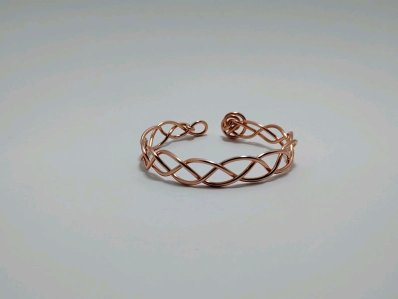 May include: A copper-colored open cuff bracelet with an intricate woven design. The bracelet features a series of intertwined loops, creating a delicate and stylish accessory. The open design allows for easy wear and adjustment. Jewelry.