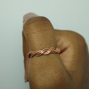 May include: A copper-colored ring with a woven design. The ring is made of thin, intertwined metal strands, creating a textured, braided appearance. The ring is worn on a finger, showcasing its intricate craftsmanship and simple elegance.