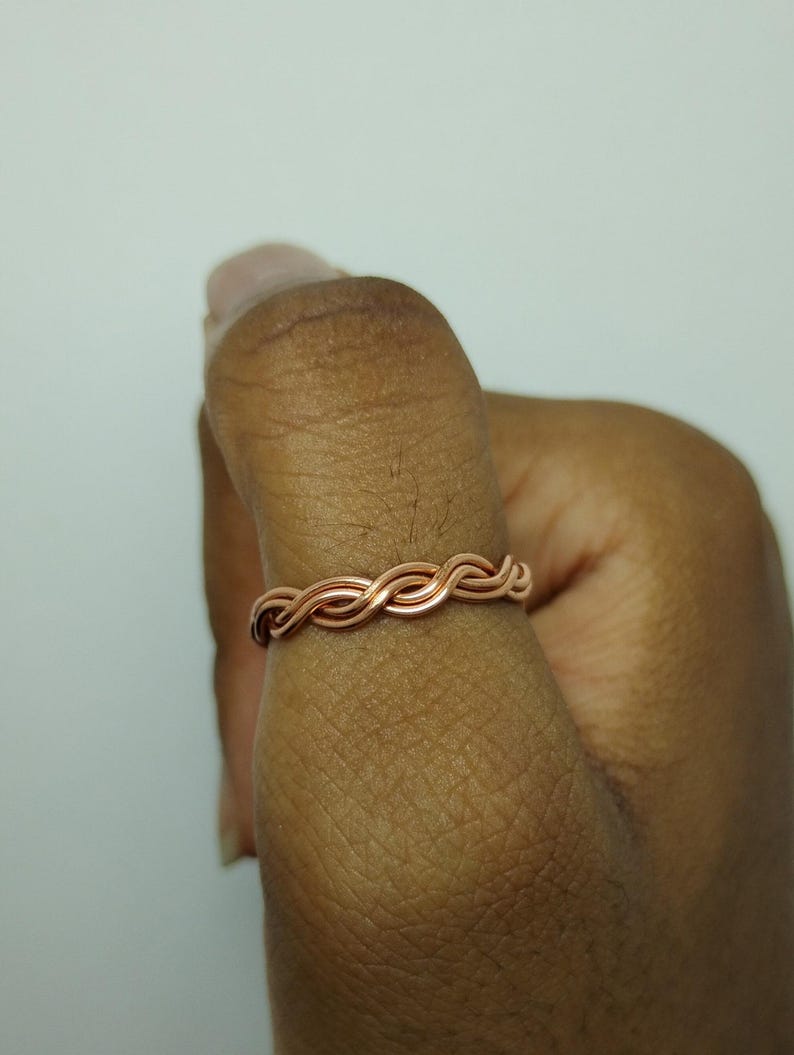 May include: A copper-colored ring with a woven design. The ring is made of thin, intertwined metal strands, creating a textured, braided appearance. The ring is worn on a finger, showcasing its intricate craftsmanship and simple elegance.