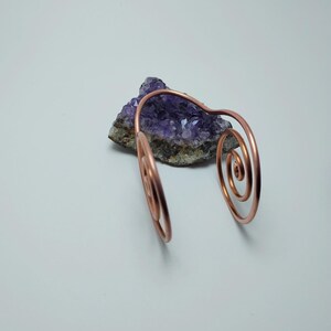 K&ouml;nnte beinhalten: Ein kupferfarbenes Armband mit Spiralmustern. Das Armband liegt auf einem lila Amethystkristall. Der Hintergrund ist eine schlichte wei&szlig;e Oberfl&auml;che. Das Armband hat zwei spiralf&ouml;rmige Enden.