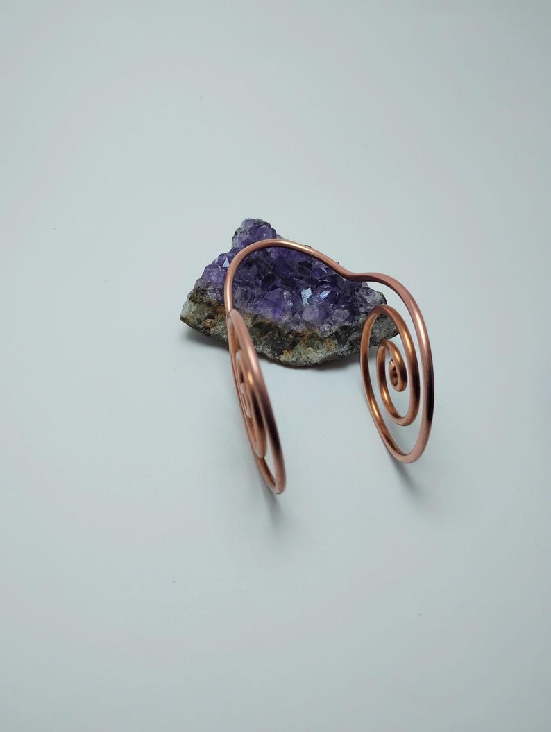 K&ouml;nnte beinhalten: Ein kupferfarbenes Armband mit Spiralmustern. Das Armband liegt auf einem lila Amethystkristall. Der Hintergrund ist eine schlichte wei&szlig;e Oberfl&auml;che. Das Armband hat zwei spiralf&ouml;rmige Enden.