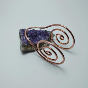 K&ouml;nnte beinhalten: Ein kupferfarbener Armreif mit Spiraldesign. Der Armreif liegt auf einem St&uuml;ck lila Amethyst. Das Metall ist in eine gebogene Form mit zwei spiralf&ouml;rmigen Enden gebogen. Der Hintergrund ist schlicht wei&szlig;.