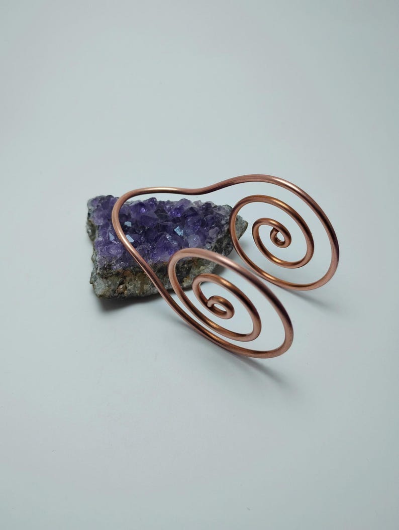K&ouml;nnte beinhalten: Ein kupferfarbener Armreif mit Spiraldesign. Der Armreif liegt auf einem St&uuml;ck lila Amethyst. Das Metall ist in eine gebogene Form mit zwei spiralf&ouml;rmigen Enden gebogen. Der Hintergrund ist schlicht wei&szlig;.