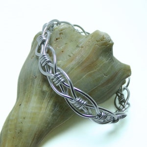 Peut inclure: Un bracelet en fil d'argent avec un design tressé, présenté sur un coquillage.
