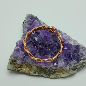 May include: A copper-colored, twisted wire bracelet rests on a purple amethyst geode. The bracelet has an open design and a textured appearance. The amethyst geode has a rough, natural surface.