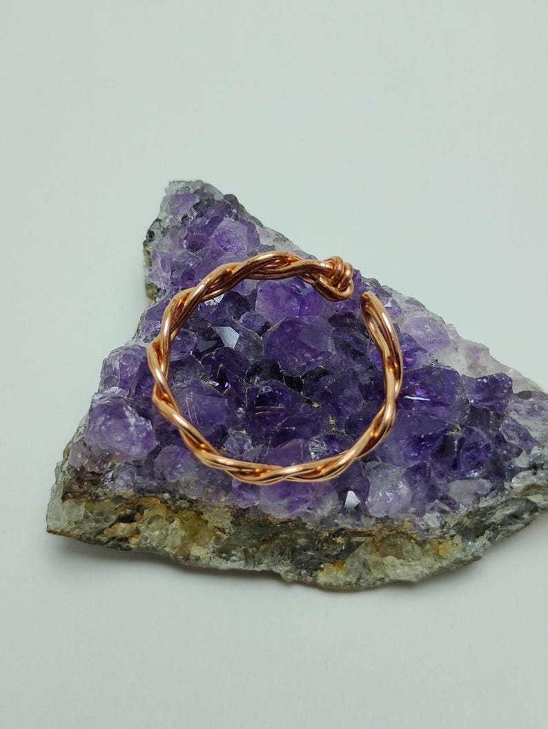 May include: A copper-colored, twisted wire bracelet rests on a purple amethyst geode. The bracelet has an open design and a textured appearance. The amethyst geode has a rough, natural surface.