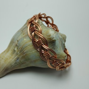 May include: A copper-colored bracelet with a woven design. The bracelet is made of intertwined wire, creating a textured, openwork pattern. The jewelry is displayed on a light-colored, textured surface.