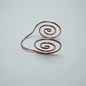 K&ouml;nnte beinhalten: Ein kupferfarbenes Drahtarmband mit zwei Spiralmustern. Das Armband hat ein schlichtes, elegantes Design, wobei die Spiralen in entgegengesetzte Richtungen zeigen. Das Metall hat eine gl&auml;nzende Oberfl&auml;che, vor wei&szlig;em Hintergrund.