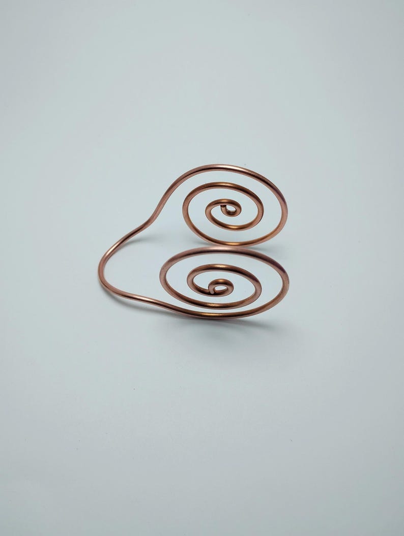 K&ouml;nnte beinhalten: Ein kupferfarbenes Drahtarmband mit zwei Spiralmustern. Das Armband hat ein schlichtes, elegantes Design, wobei die Spiralen in entgegengesetzte Richtungen zeigen. Das Metall hat eine gl&auml;nzende Oberfl&auml;che, vor wei&szlig;em Hintergrund.