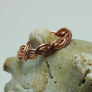 May include: A copper-coloured ring with a braided design. The ring is made of intertwined metal strands, creating a textured, woven appearance. The ring is displayed on a light-coloured, textured surface.